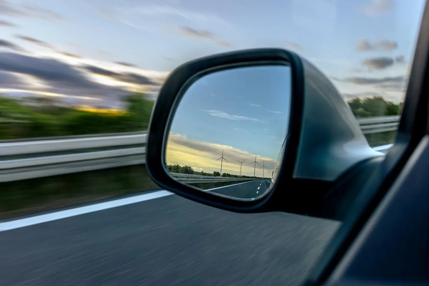 Боковое зеркало автомобиля, ветряные турбины на горизонте, скорость и устойчивое развитие.